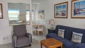 A living room with a grey armchair, a blue sofa, and a wooden coffee table. There is a dining area with a white table and chairs, and a kitchen with cabinets and a window. - Breakaway Inn (Lauderdale-by-the-Sea)