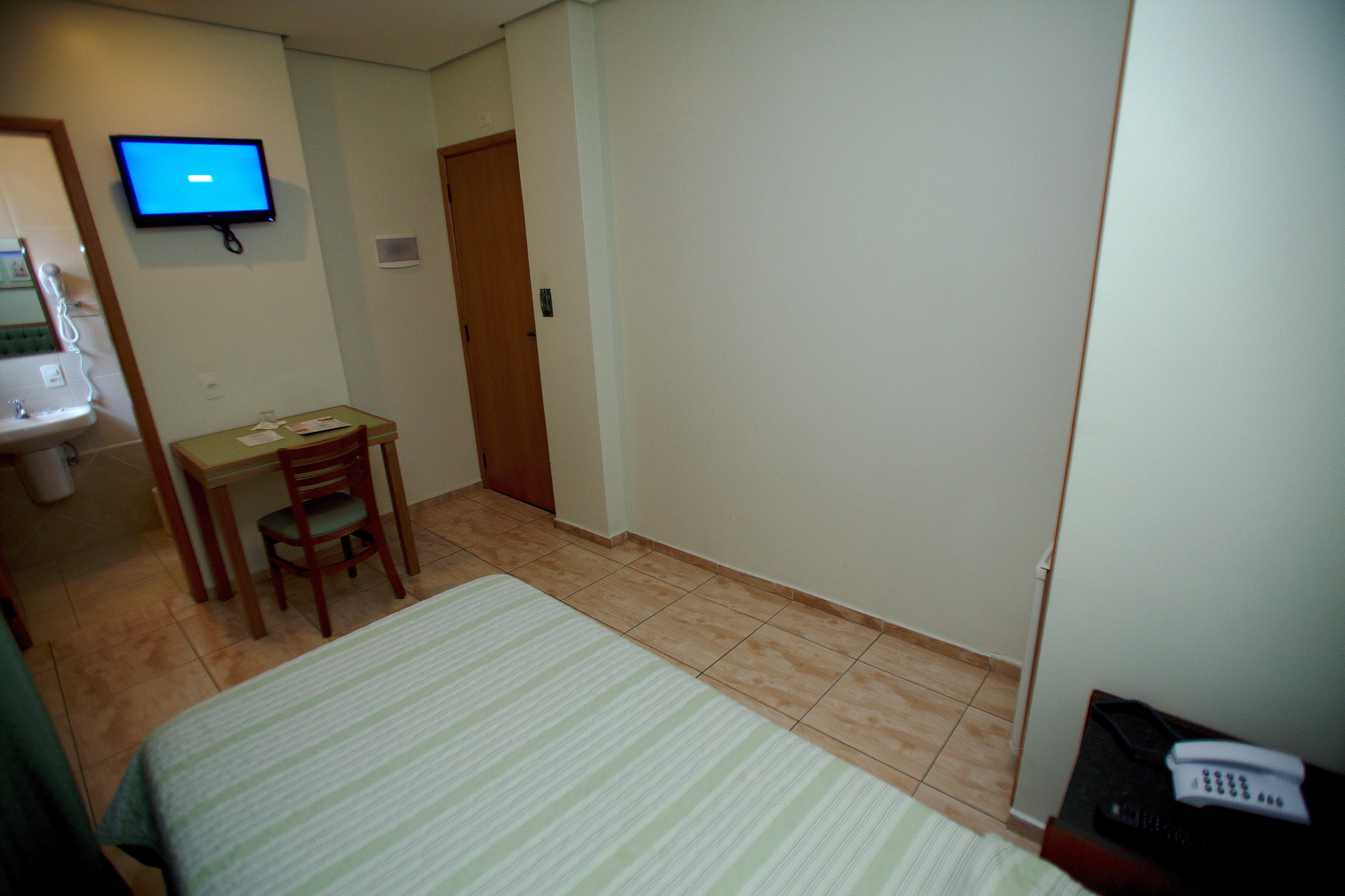 Una habitación de hotel pequeña con una cama, un escritorio, una silla, un televisor instalado en la pared y un baño con lavabo y espejo.