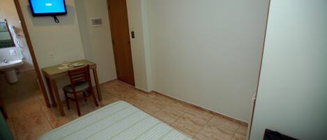 Una habitación de hotel pequeña con una cama, un escritorio, una silla, un televisor instalado en la pared y un baño con lavabo y espejo.