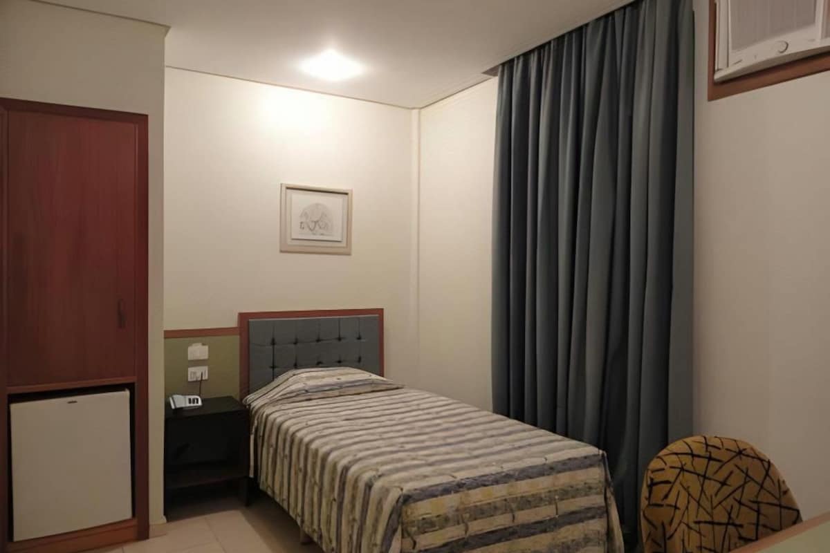 Un dormitorio con cama, mesita de noche, escritorio, armario y ventana con cortinas.
