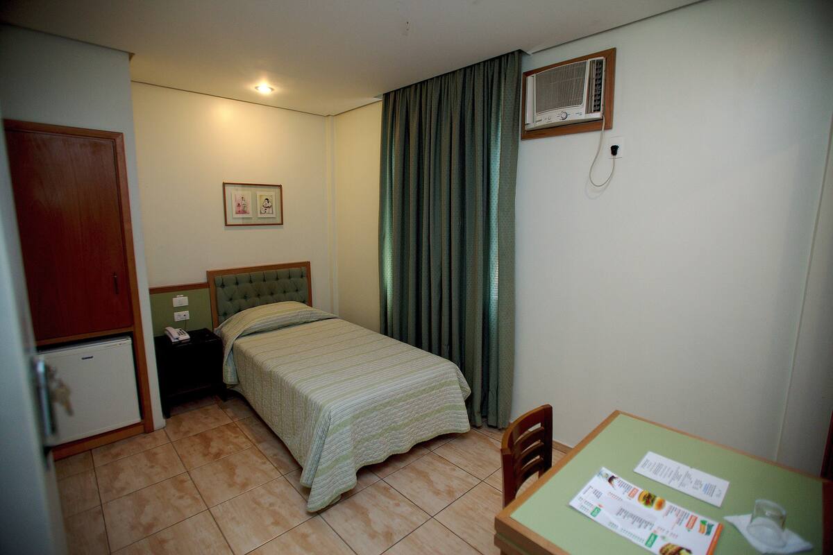 Une chambre d’hôtel comprenant un lit, un petit bureau, une chaise, un réfrigérateur, une table de chevet, une fenêtre avec des rideaux et un climatiseur fixé au mur.