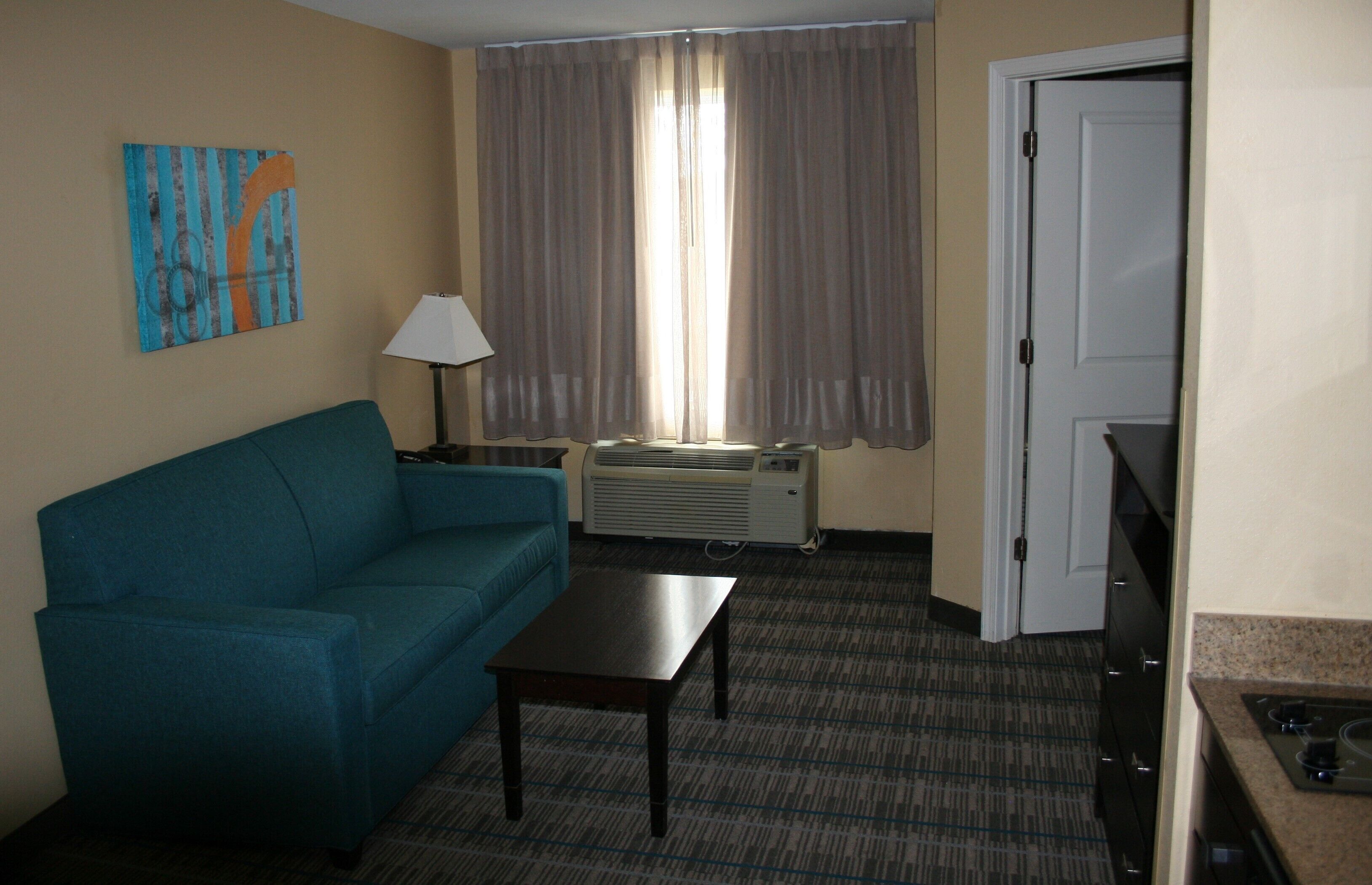 A hotel room with a blue sofa, a small table, a lamp, an air conditioning unit, and a kitchen area with a stove.