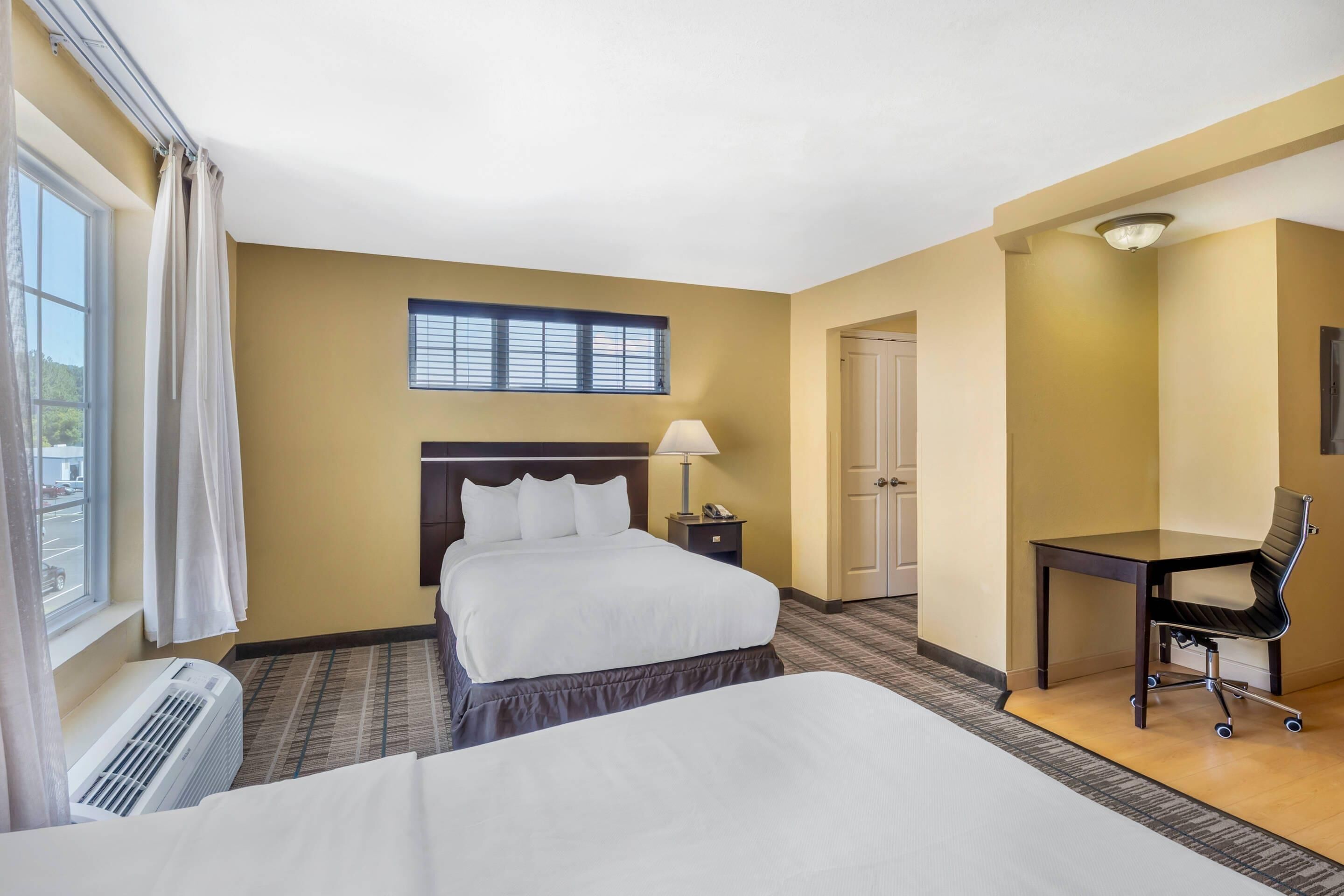 Una habitación de hotel con cama, escritorio, silla, lámpara y ventana con cortinas.