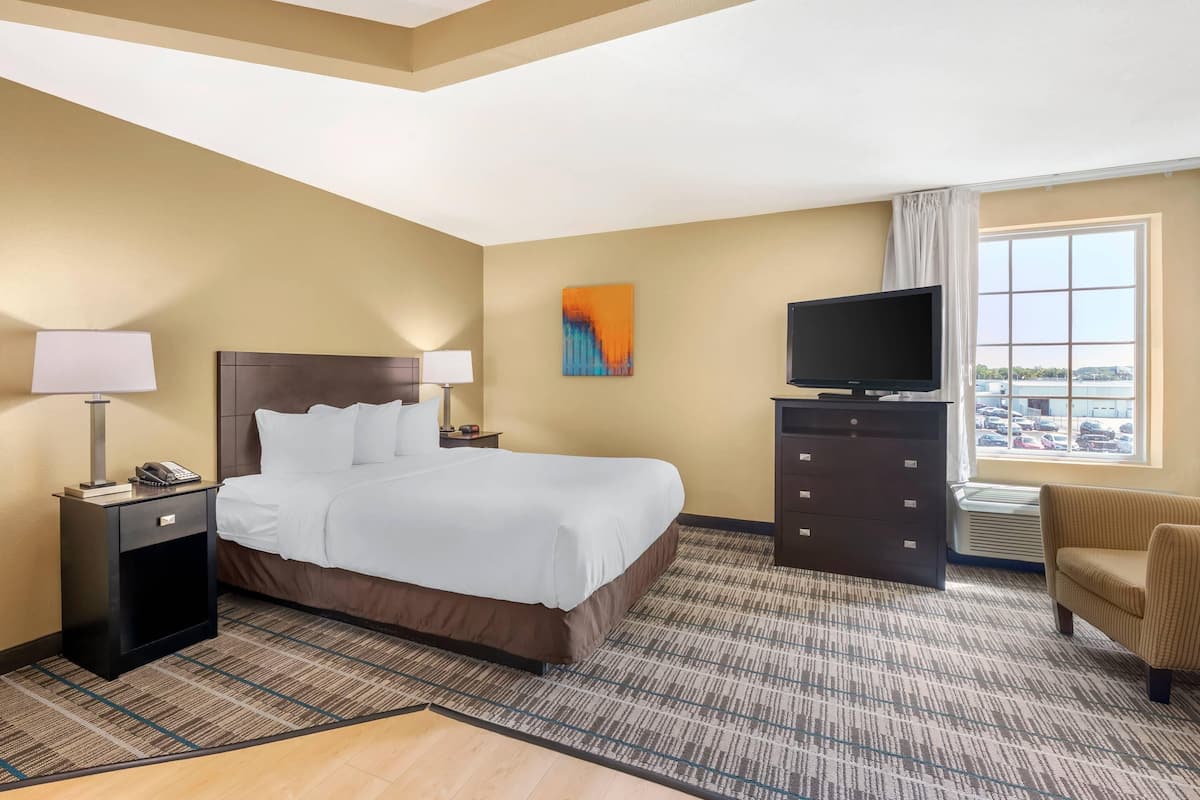 A hotel room with a bed, a TV, a desk with a lamp, a chair, and a window with a view of the parking lot.