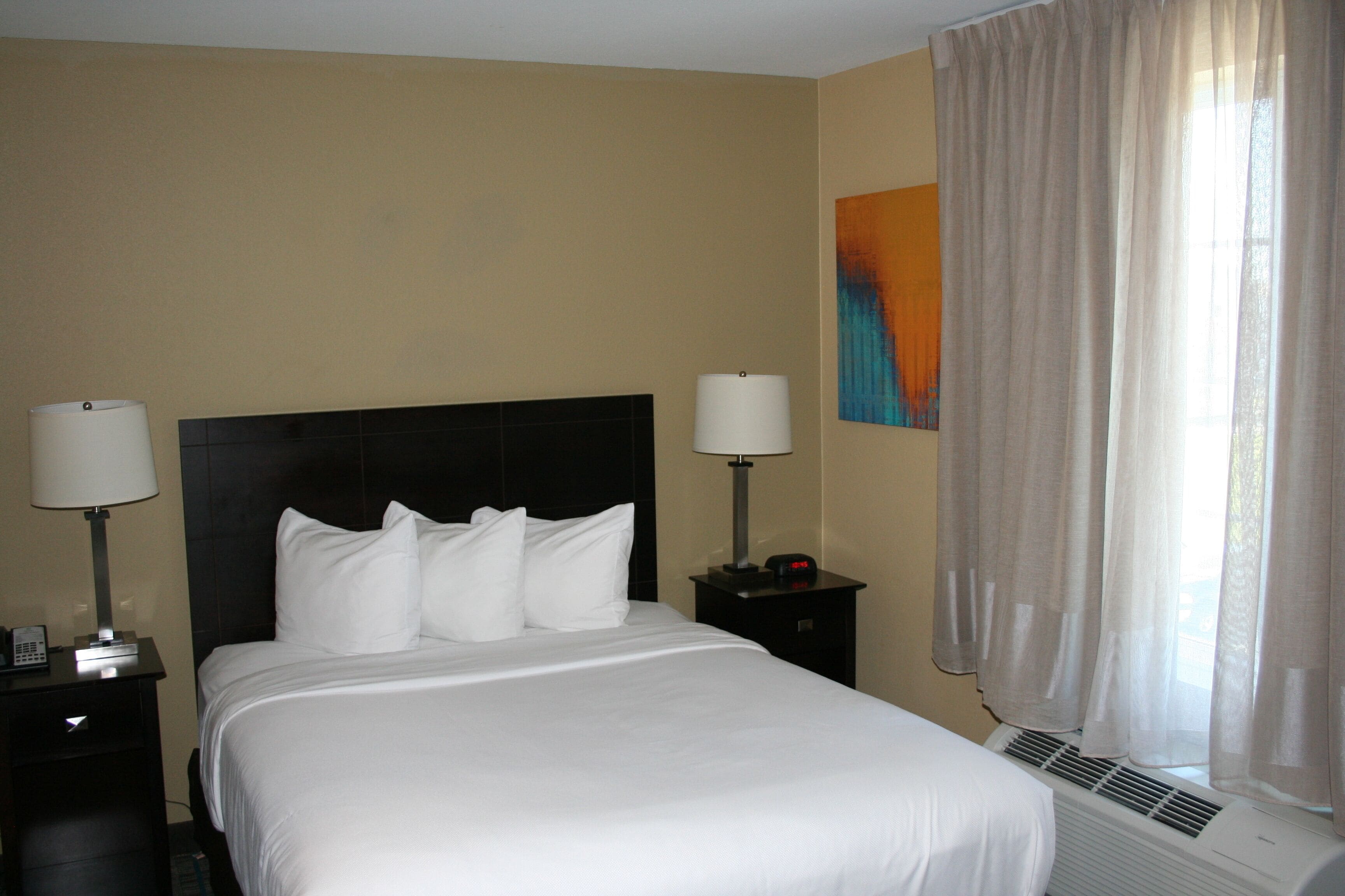 A hotel room with a bed, two bedside tables, a nightstand, a wall-mounted air conditioner, and a window with curtains.