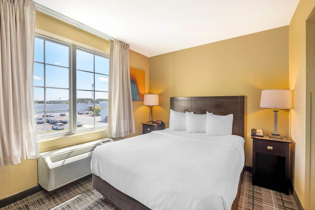 A hotel room with a large bed, two bedside tables with lamps, a window with curtains, and a view of a parking lot.
