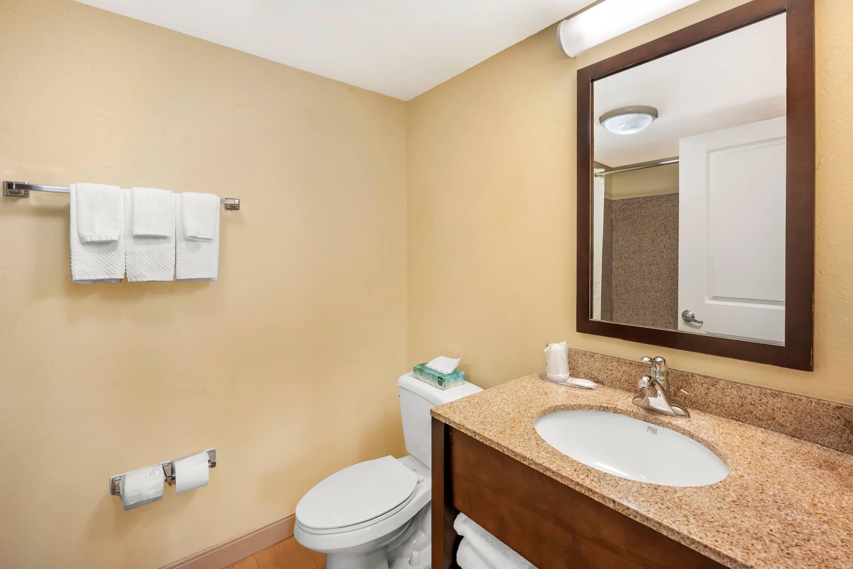 A bathroom with a beige wall, a white toilet, a sink with a mirror above it, and a towel rack with three towels.