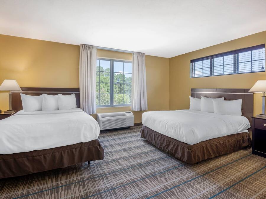 A hotel room with two beds, a large window with curtains, an air conditioning unit, and a view of greenery outside.