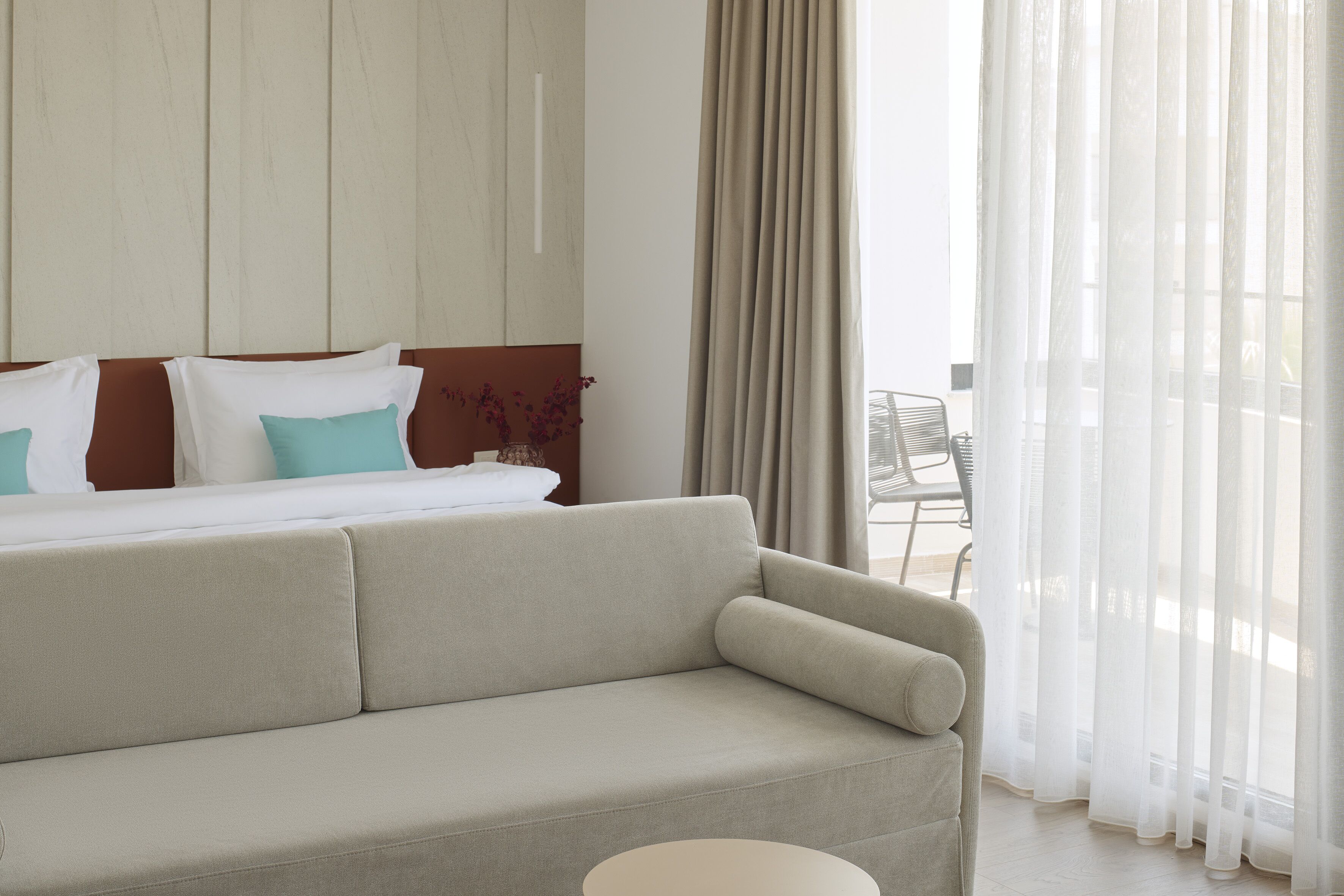 Een hotelkamer met een bed, een bank, een klein tafeltje en een raam met dunne gordijnen.