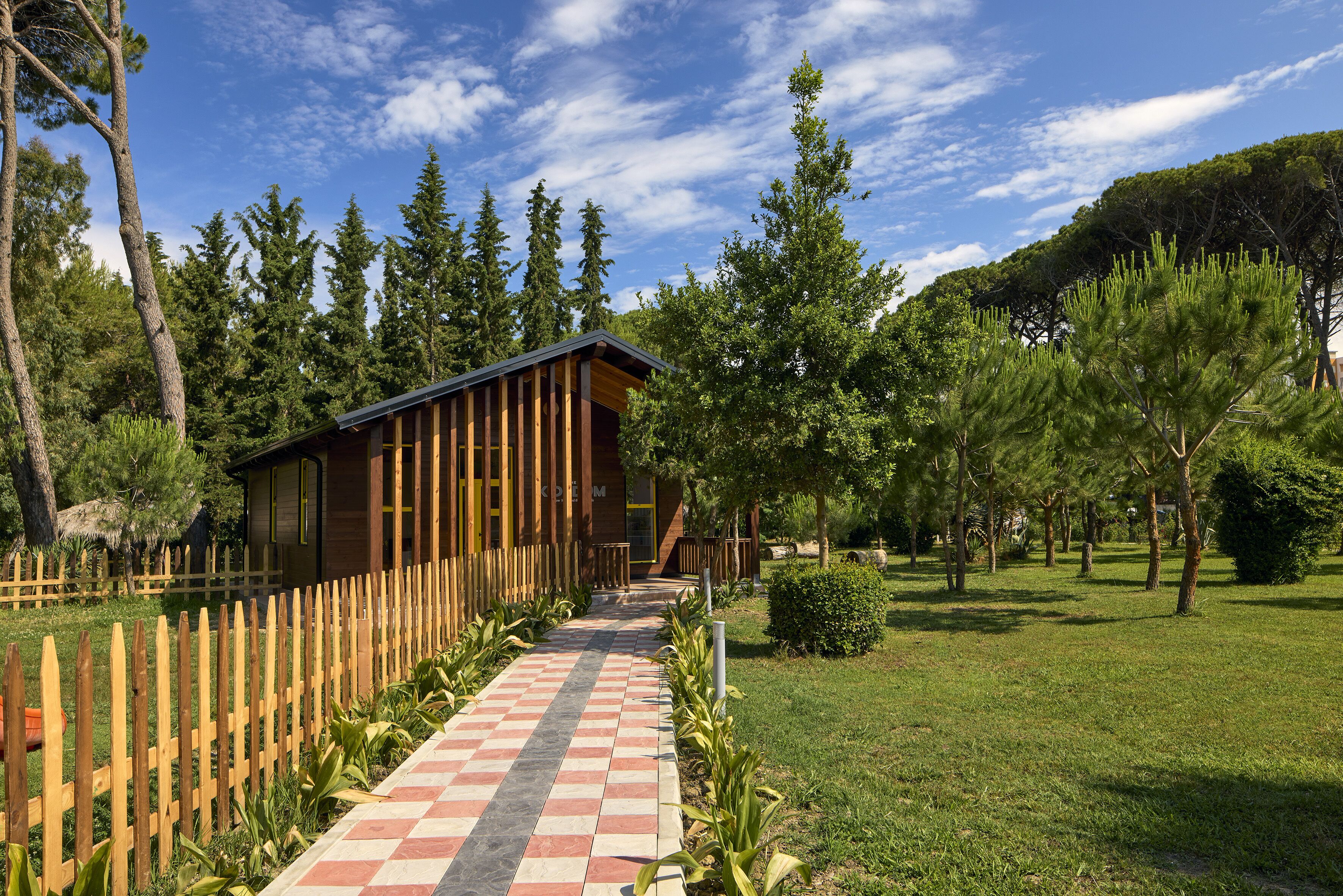A wooden cabin with a checkered pathway leading to it, surrounded by trees and a wooden fence.