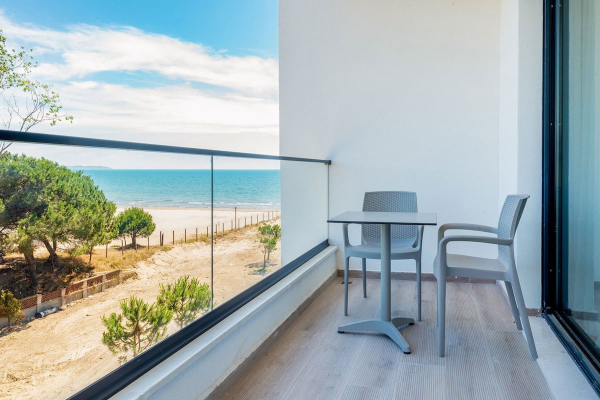 A balcony with a view of the beach, a table, and two chairs.