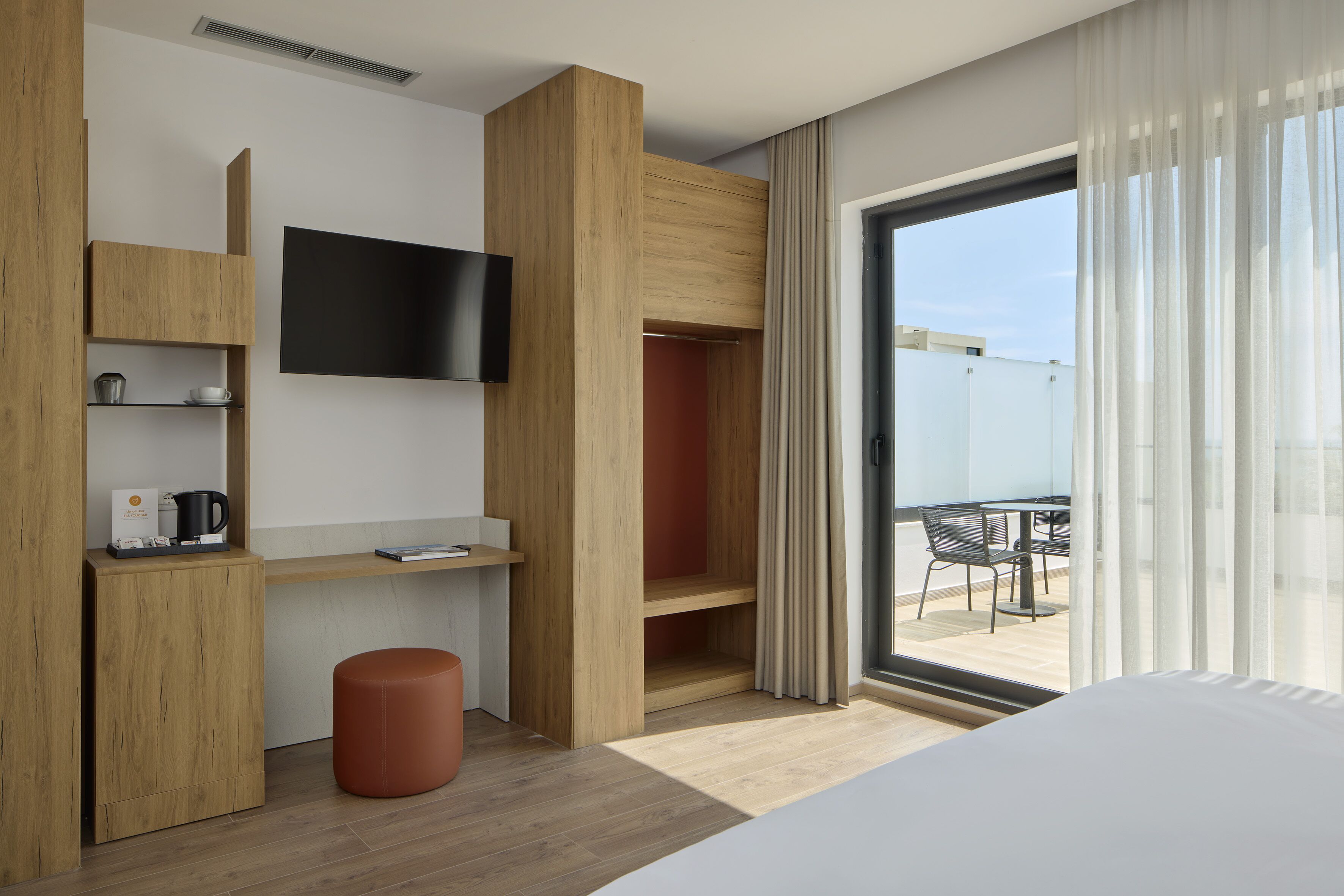 A modern hotel room with a wooden wardrobe, a flat-screen TV, a small table with a coffee maker, and a view of the outdoors through a large window.