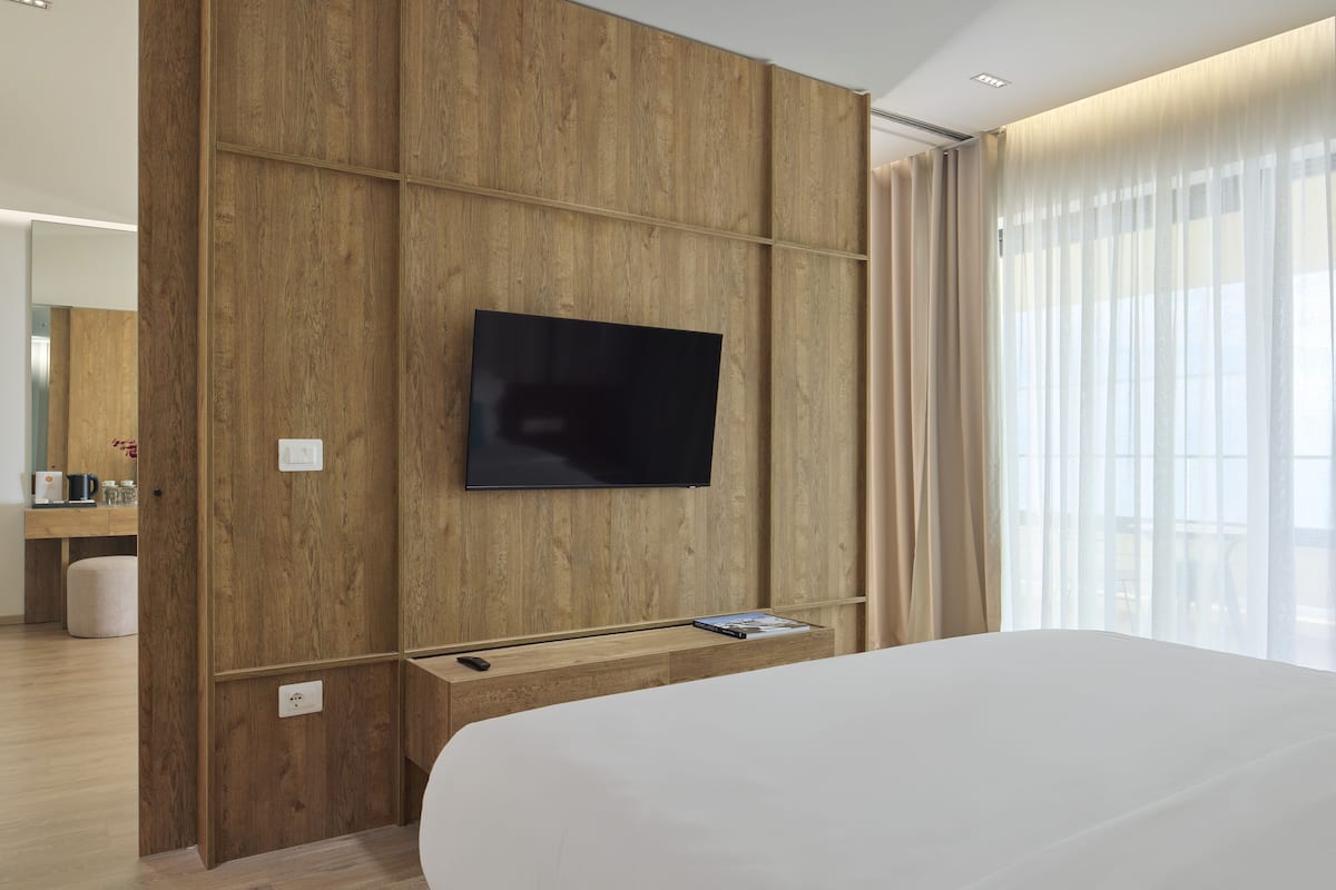 A modern hotel room with a large bed, a mounted TV, wooden paneling, and a view through sheer curtains.