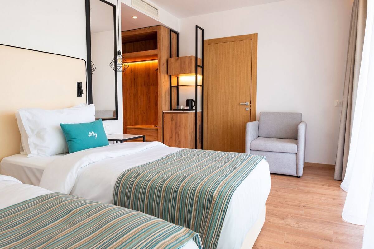A hotel room with two beds, a grey armchair, a wooden wardrobe, a small table with a coffee machine, and a mirror.
