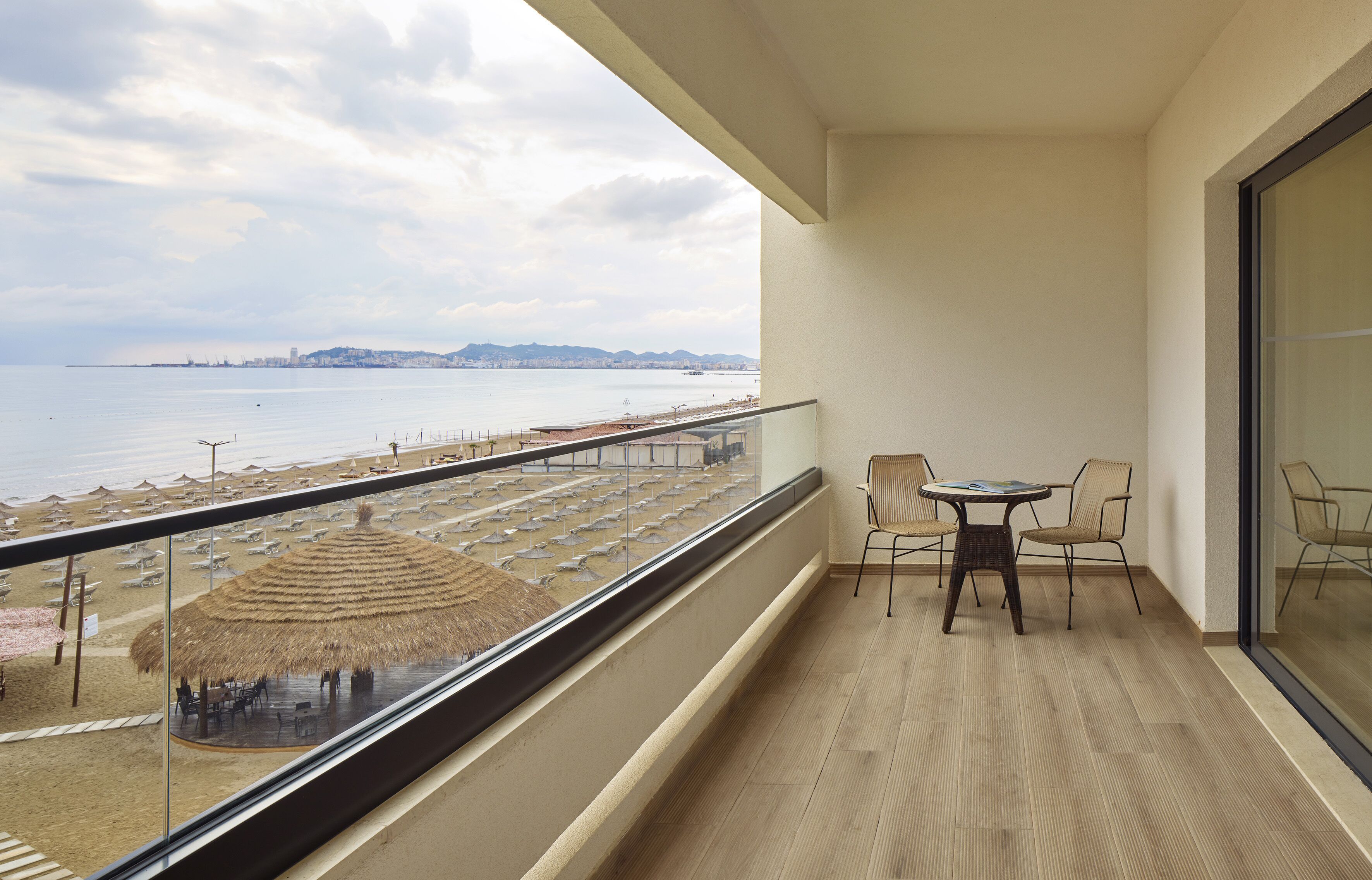 A balcony with a view of the beach, a table with chairs, and a thatched umbrella.