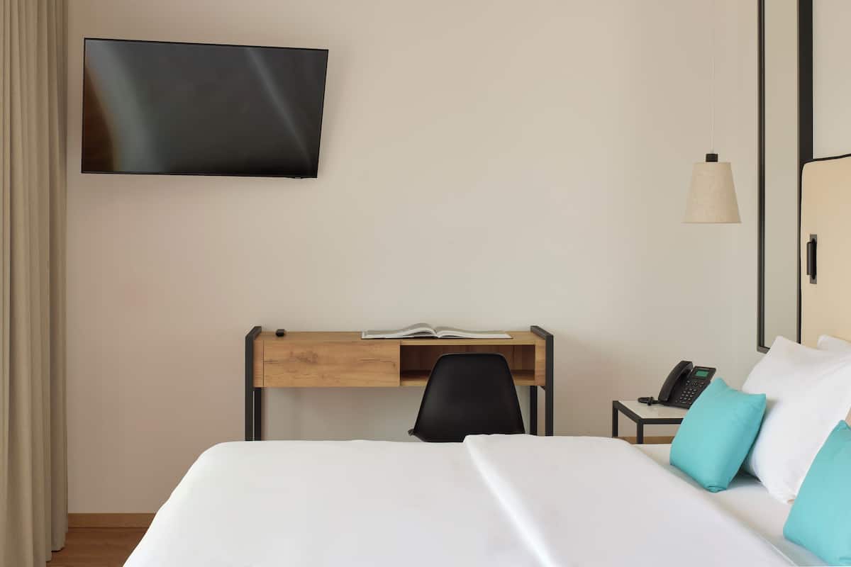 A hotel room with a bed, a desk with a chair, a television mounted on the wall, a bedside table with a phone, and a hanging light fixture.