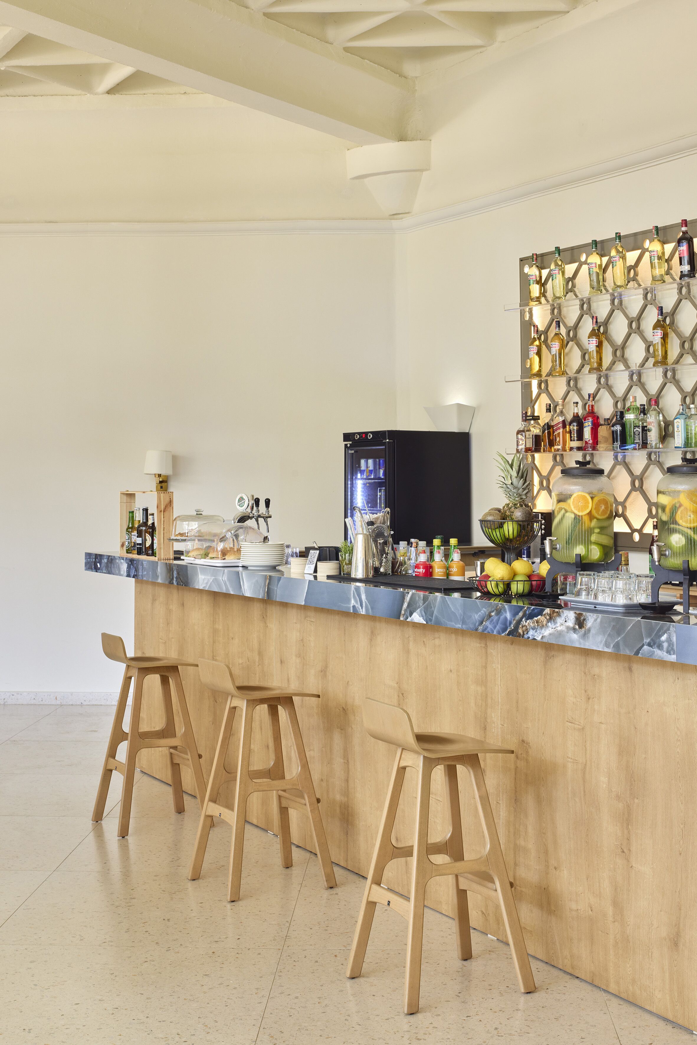 A bar area with a marble countertop, wooden stools, a fruit display, and a wall-mounted liquor shelf.