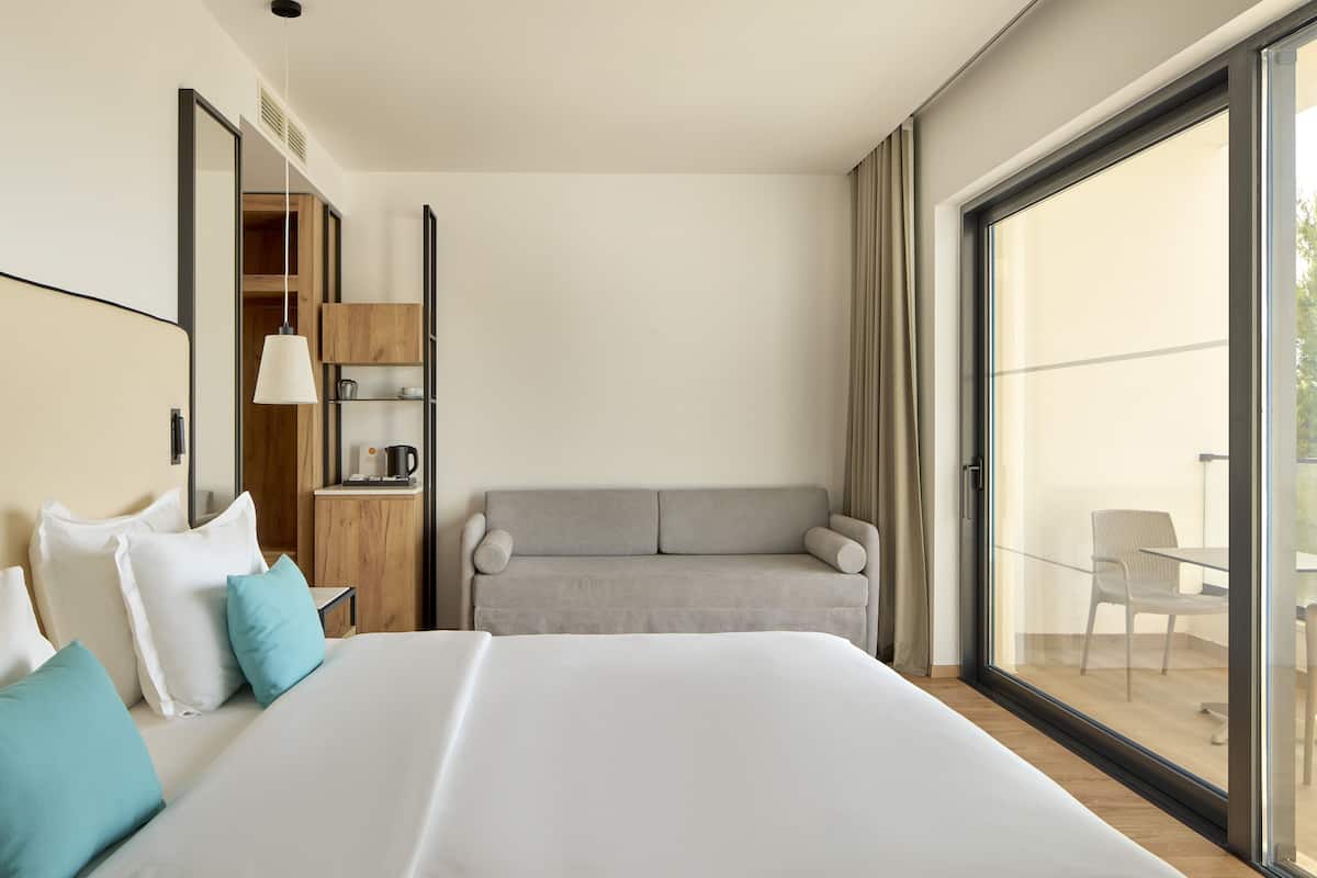 A modern hotel room with a large bed, a sofa, a dining area with a table and chairs, and a sliding glass door to the outside.