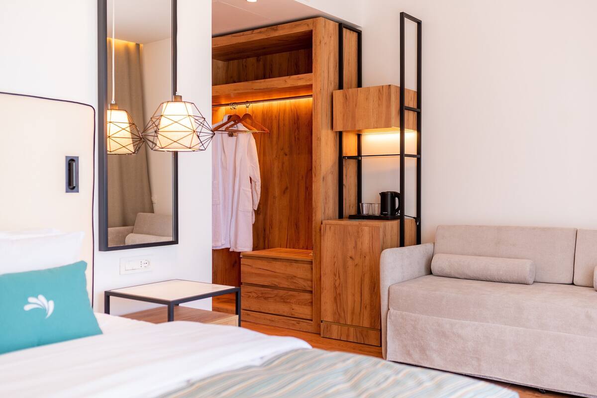A hotel room with a bed, a small table, a sofa, a wooden wardrobe, and a mirror.