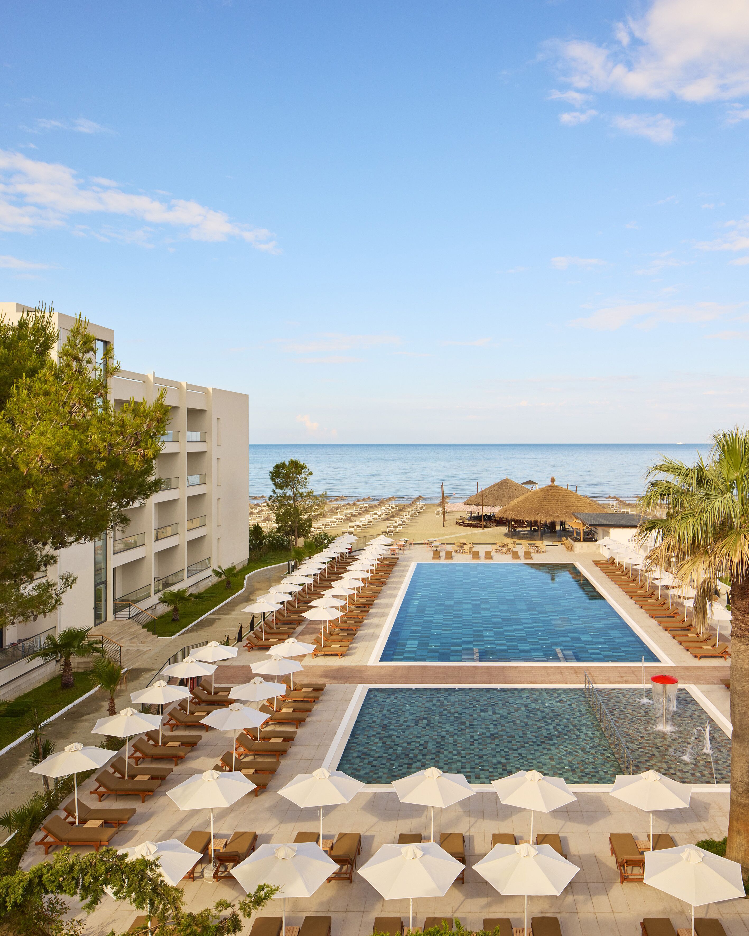 A resort pool area with lounge chairs and umbrellas, a clear view of the sea, and a modern building with balconies.
