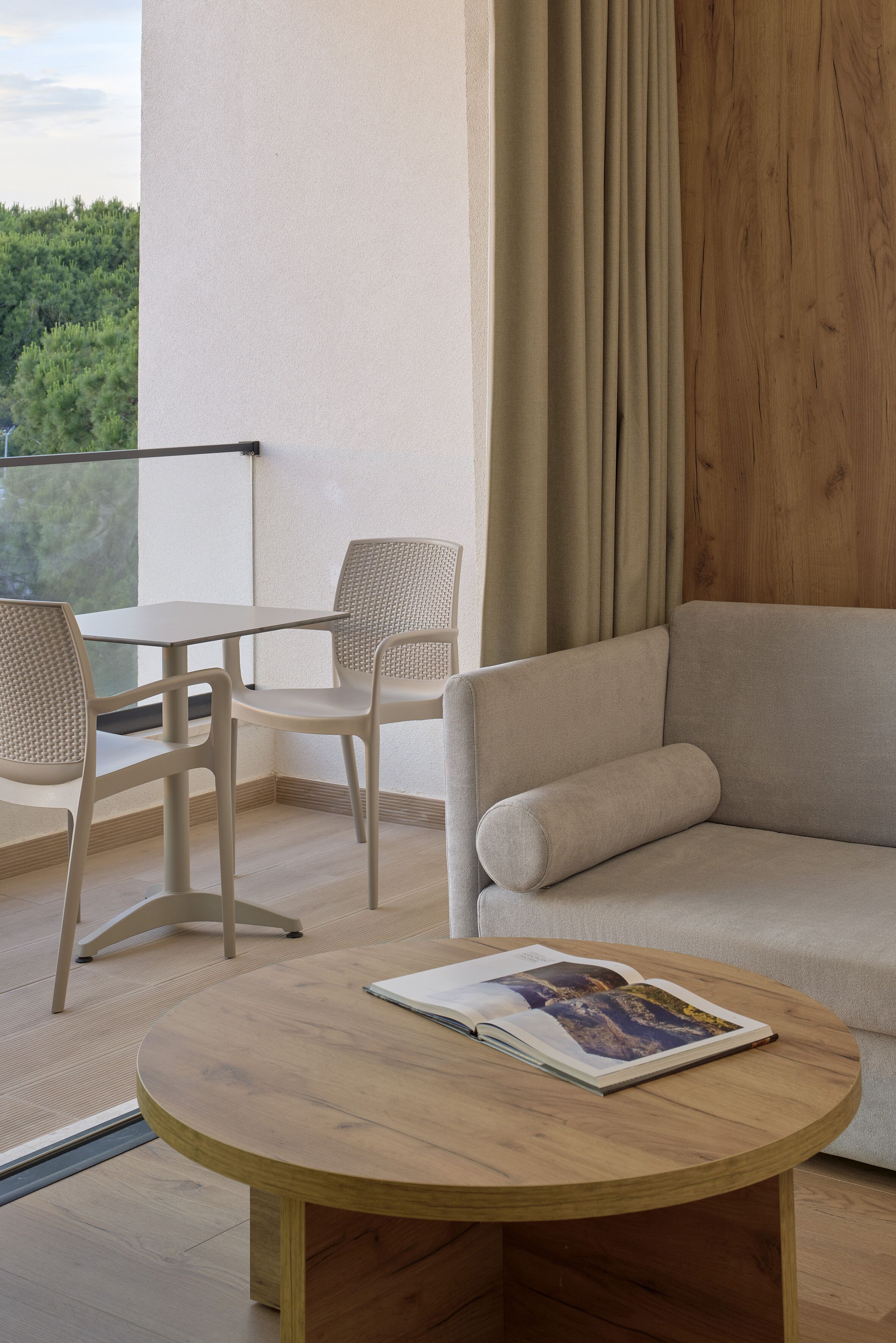 Ein modernes Zimmer mit einem runden Holztisch, einem beigen Sofa und zwei Korbstühlen. Ein Glastisch steht in der Nähe eines Fensters mit Blick auf Grünflächen.