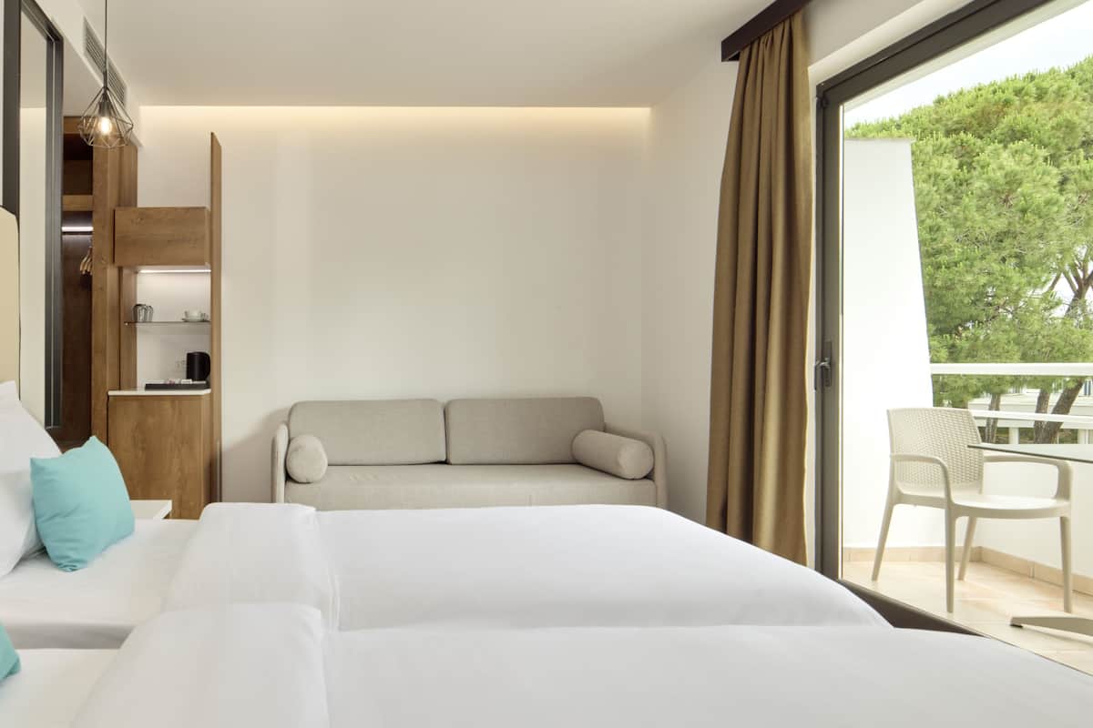 A hotel room with a bed, a sofa, a small table, a chair, and a balcony with a view of trees.