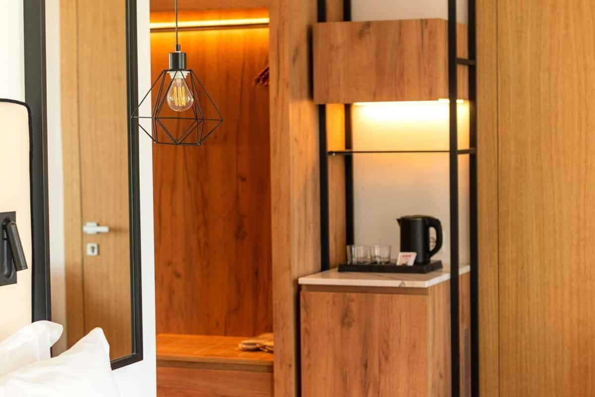 A hotel room with a bed, a wooden wardrobe, a small wooden table with a kettle, and a hanging light fixture.