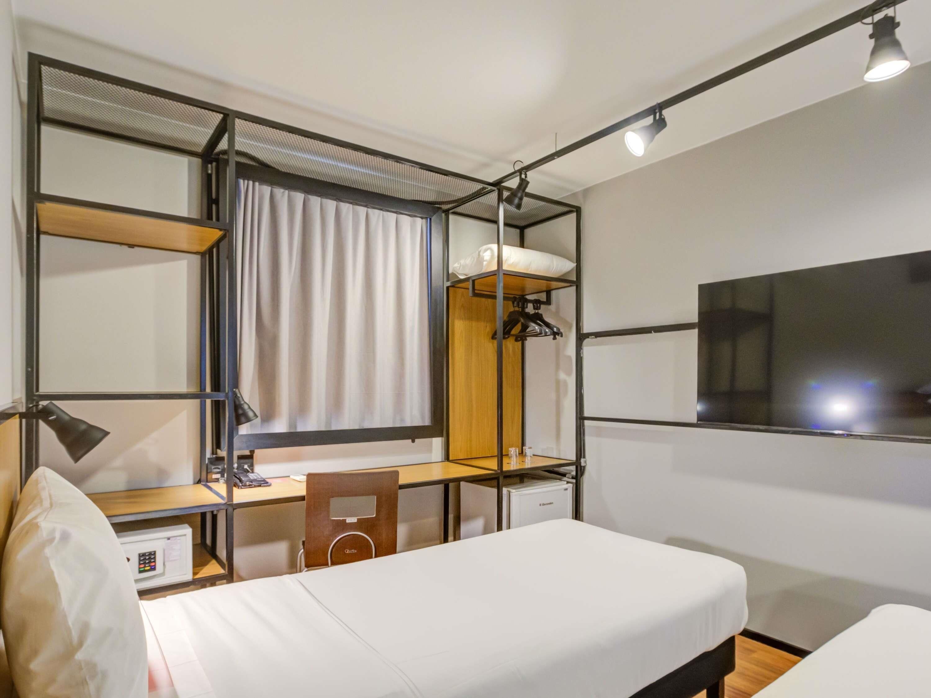 A compact hotel room with a bed, a desk with a chair, a wardrobe, a TV, and a window with curtains.