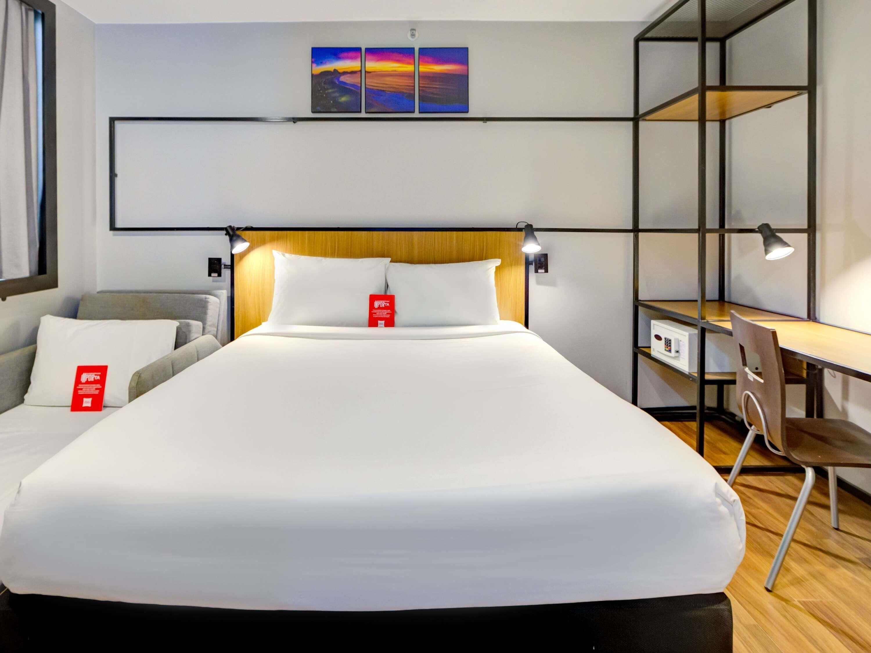 Habitación de hotel con cama, escritorio, silla, mesita de noche y estante en la pared.