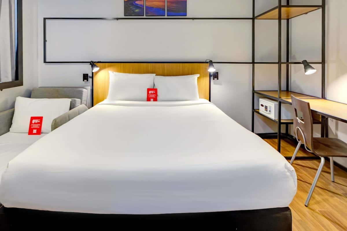 Ett hotellrum med en säng, ett skrivbord, en stol, ett nattduksbord och en hylla som är monterad på väggen.