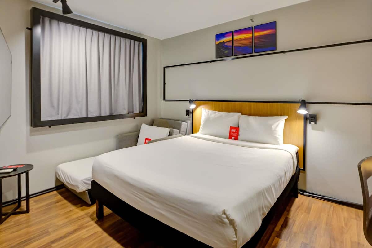 Ein Hotelzimmer mit einem großen Bett, einem Nachttisch, einem Sessel, einem Fenster mit Vorhängen und einem an der Wand hängenden Kunstwerk.