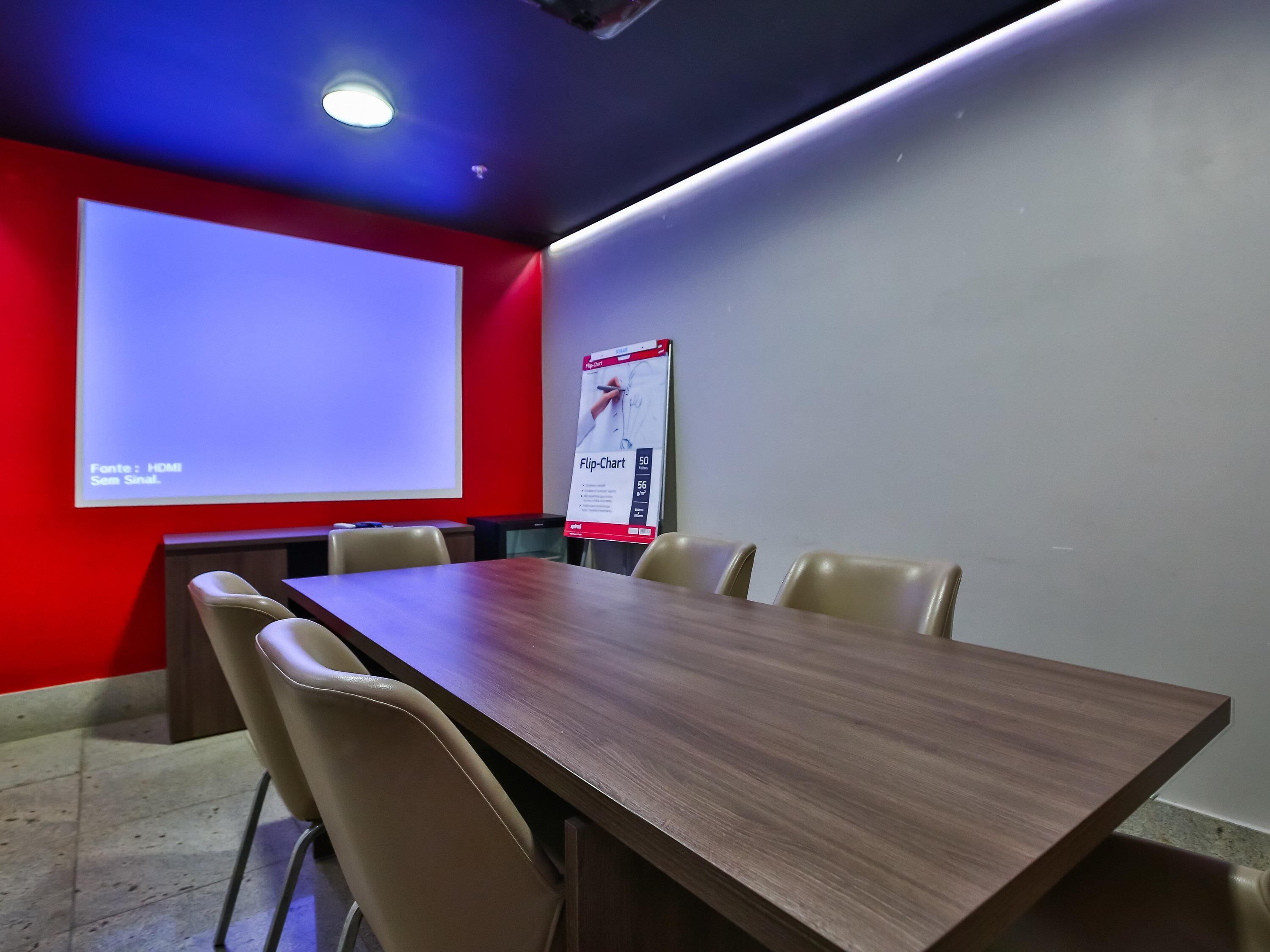 Une salle de conférence avec une grande table, des chaises de couleur beige, un écran de projection et un tableau mobile fixé au mur.