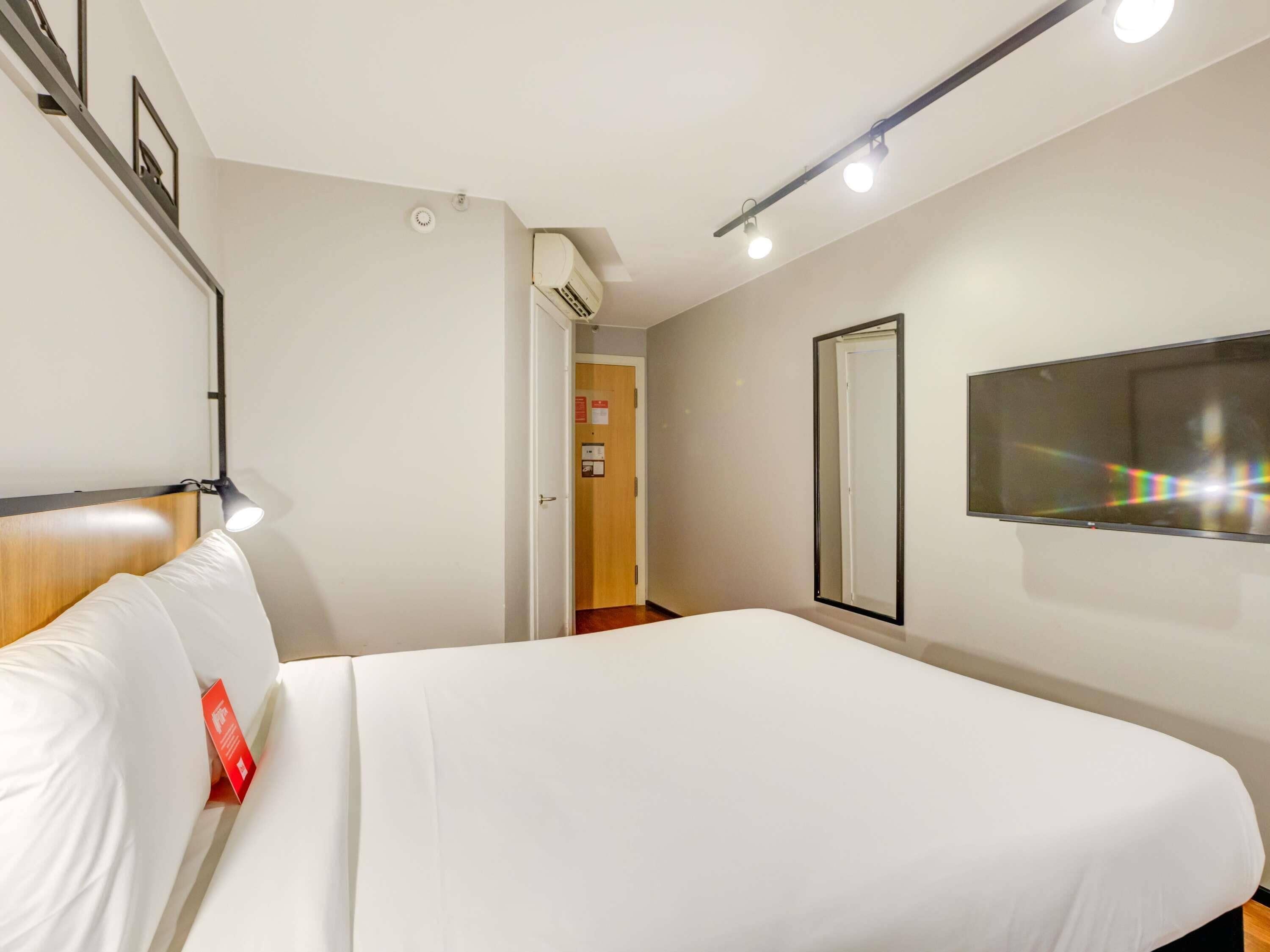 A modern hotel room with a large bed, a bedside lamp, a flat-screen TV, and an air conditioning unit.