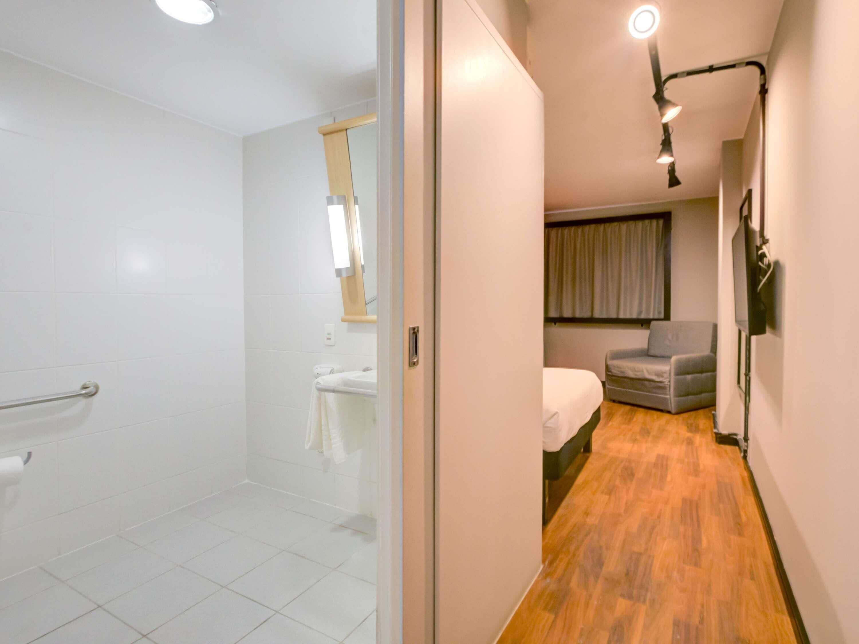 Un baño moderno con ducha, inodoro y lavamanos. Un pasillo conduce a una zona de estar con sofá y televisor.
