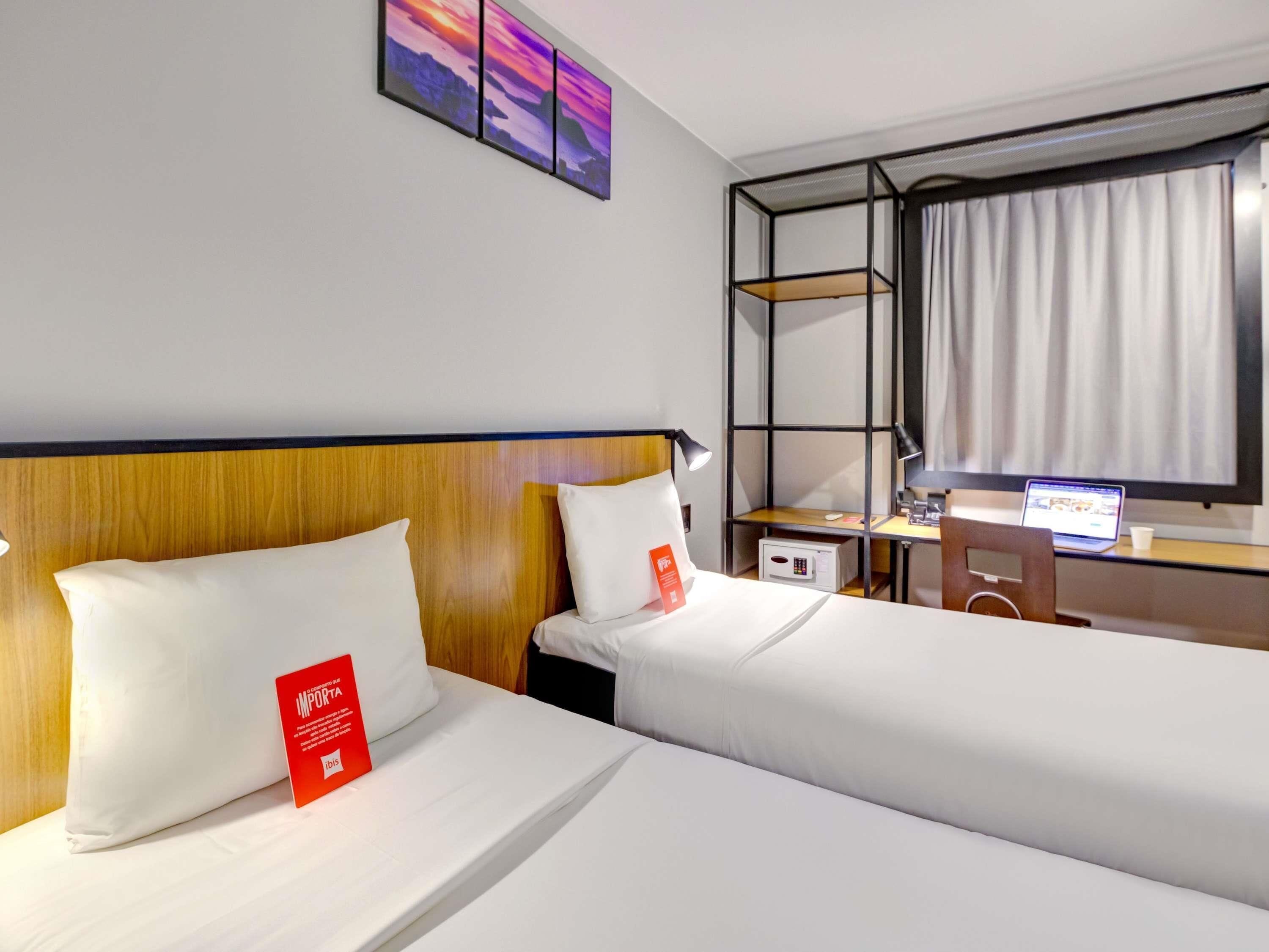 A hotel room with two beds, a desk with a laptop, a window with curtains, and a wall-mounted shelf.