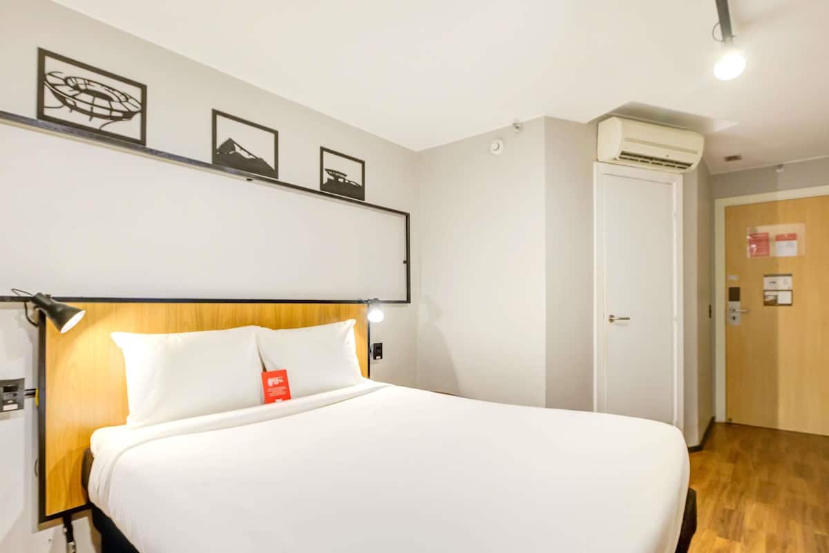 Ein Hotelzimmer mit einem Bett, einem Kopfteil, einem an der Wand befestigten Regal mit Bildern, einer Klimaanlage und einer Tür.
