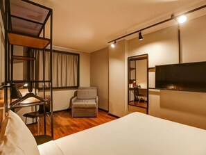 A modern hotel room with a bed, a desk, a chair, a TV, and a large mirror.