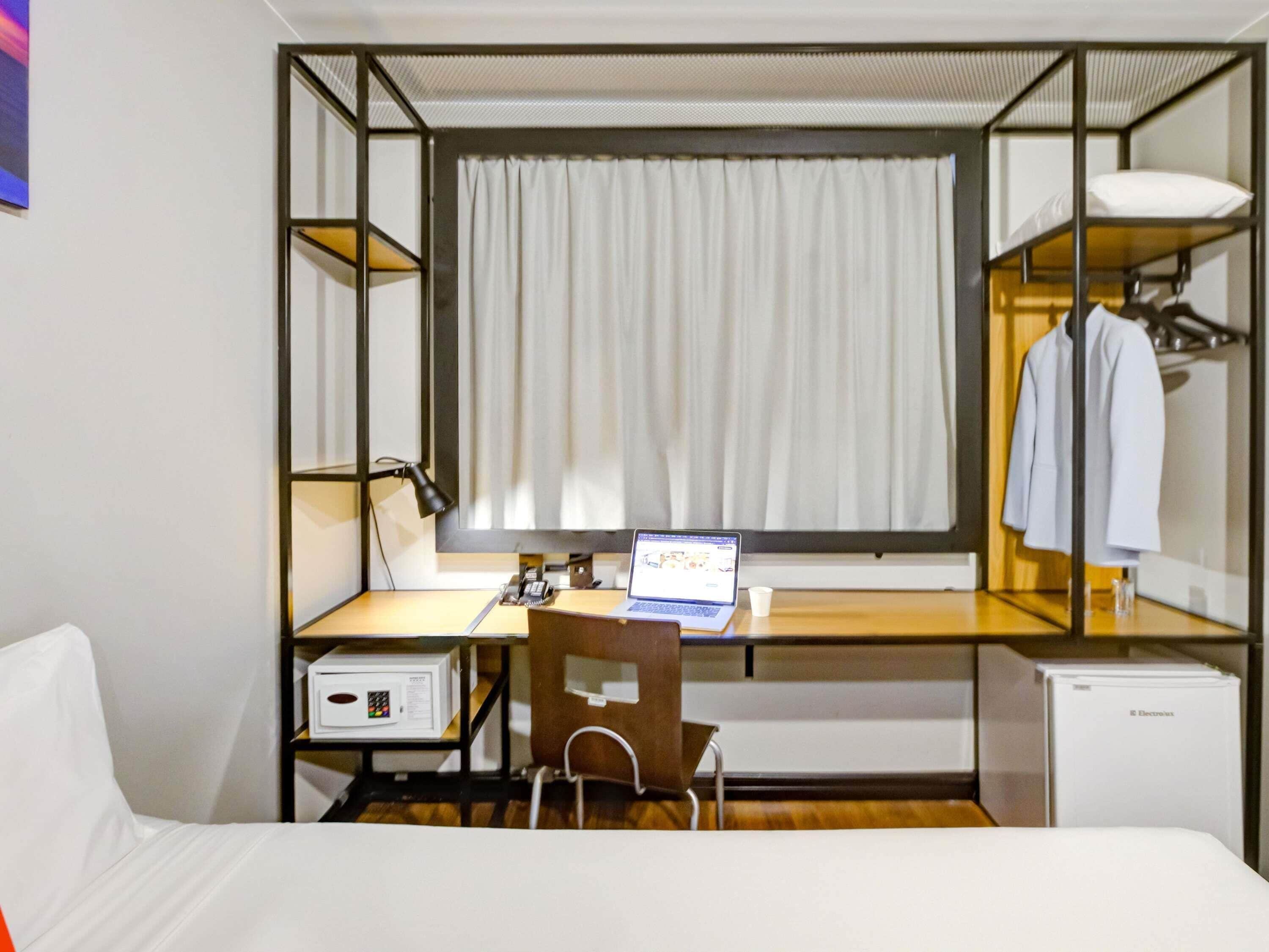 Een kleine hotelkamer met een eenpersoonsbed, een bureau met een laptop, een stoel, een kleine koelkast en een kledingkast.