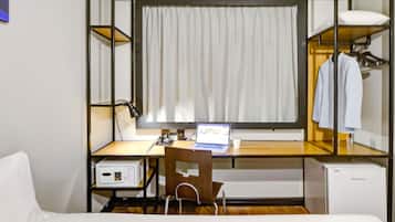 Een kleine hotelkamer met een eenpersoonsbed, een bureau met een laptop, een stoel, een kleine koelkast en een kledingkast.
