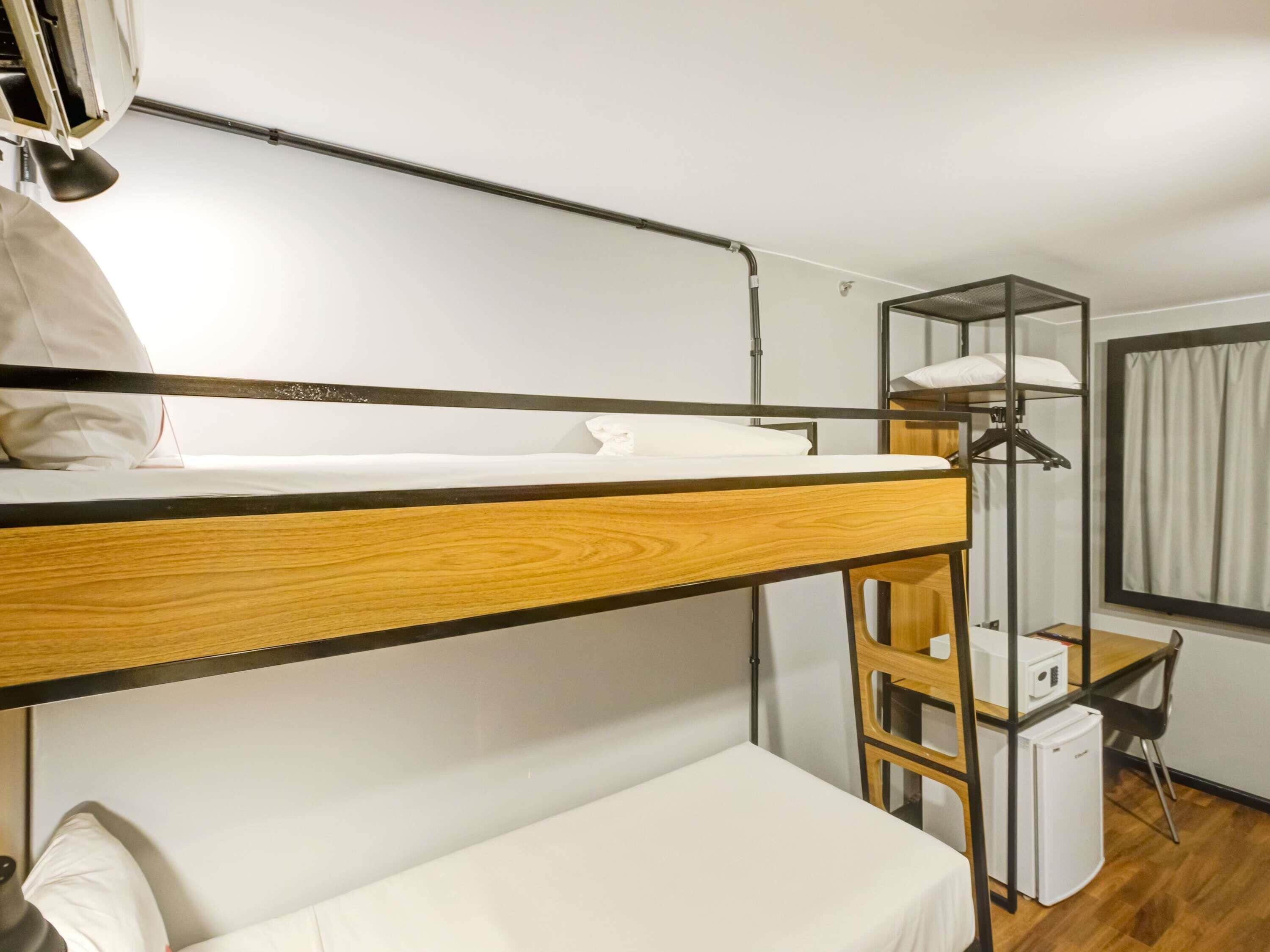 A bunk bed with a wooden frame, a desk with a chair, a small refrigerator, and a window with curtains.