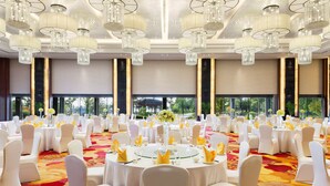 Un salón de banquetes con mesas redondas, sillas blancas, candelabros y ventanas amplias.