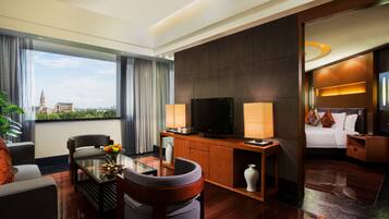 A modern hotel room with a large window, a flat-screen TV, a sofa, a dining table, and a bed with a nightstand.