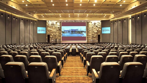 Un salón de conferencias con filas de sillas, una pantalla grande al frente y un fondo de pared de piedra.