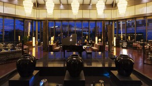 A spacious lobby with a grand piano, seating areas, and large windows offering a city view.