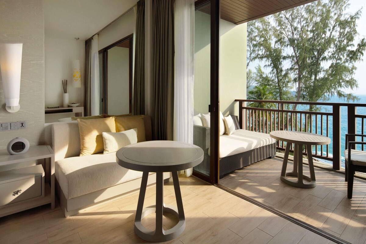 Quarto de hotel moderno com varanda, sofá, mesa redonda e vista para o mar.