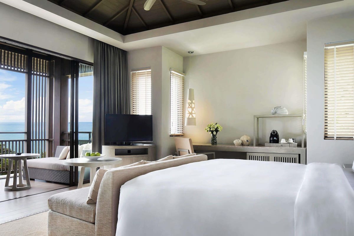 Quarto de hotel com uma cama grande, televisão, área de estar com mesa e vista para o mar.
