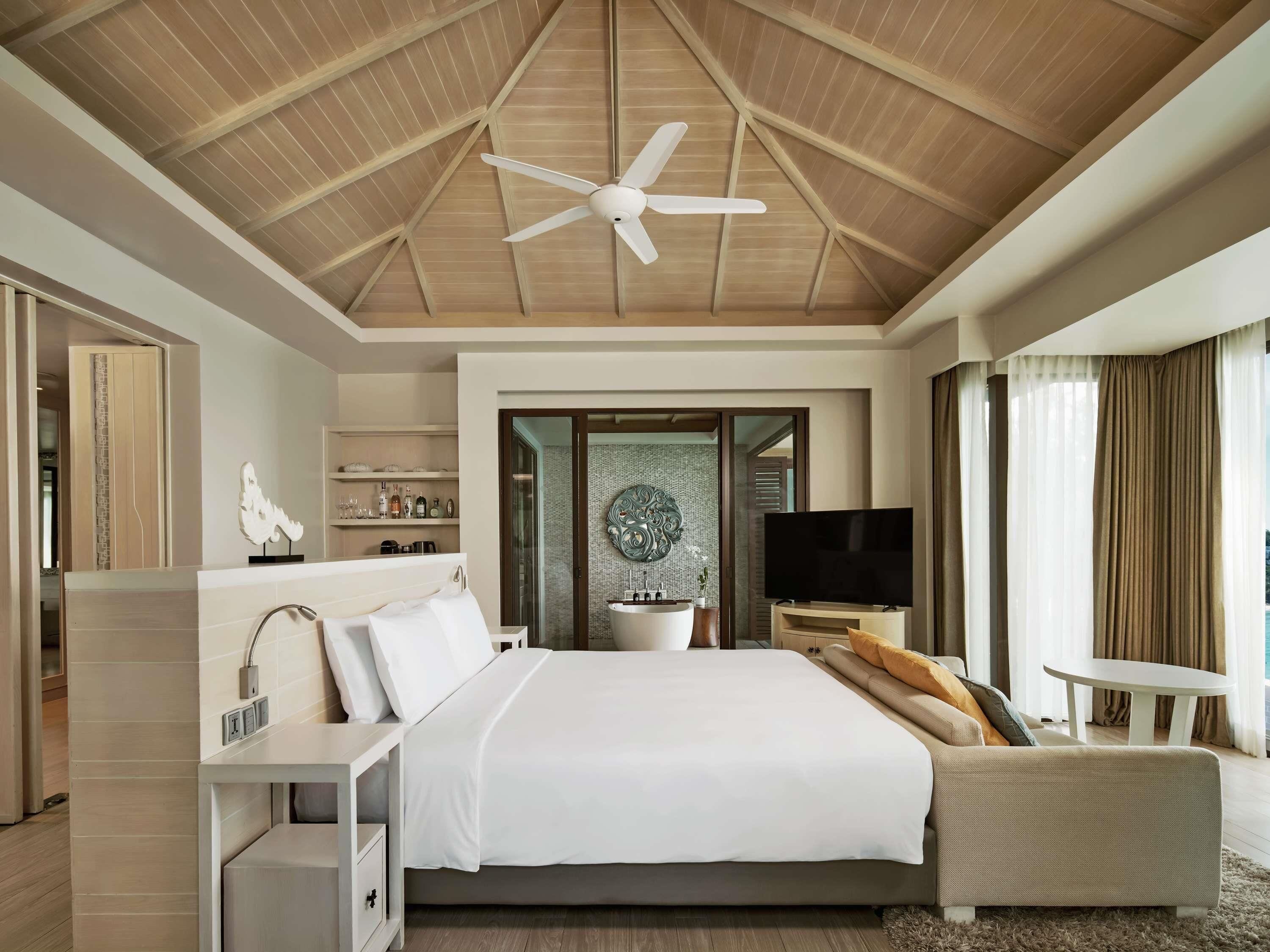 A modern hotel room with a large bed, a TV, a ceiling fan, and a view of the sea.