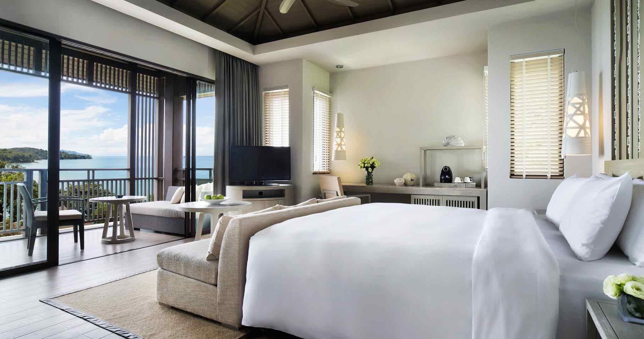 A modern hotel room with a large bed, a balcony with a view of the sea, a TV, and a small dining area.