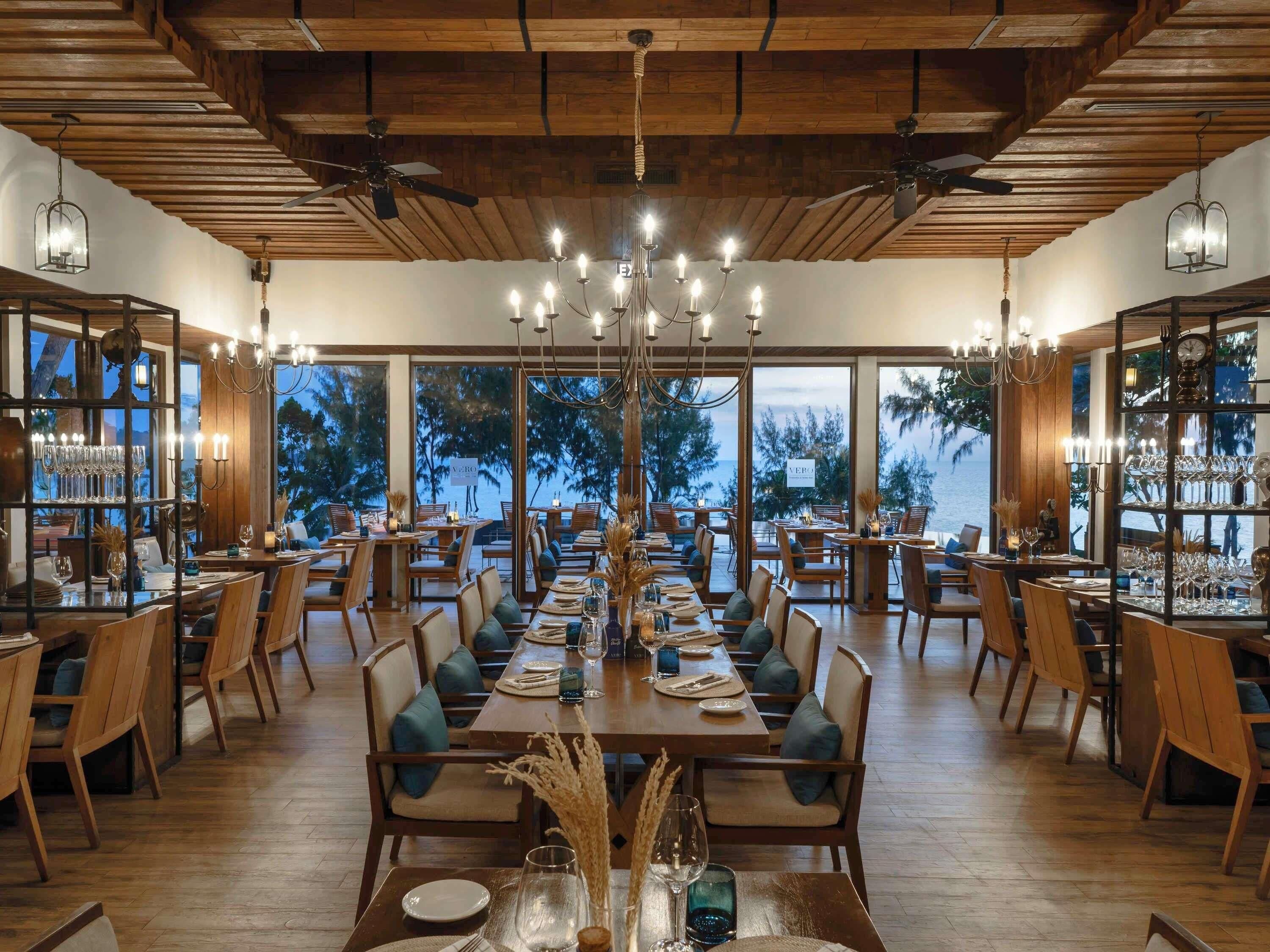 A spacious dining area with wooden tables and chairs, large windows offering a view of trees and a body of water.