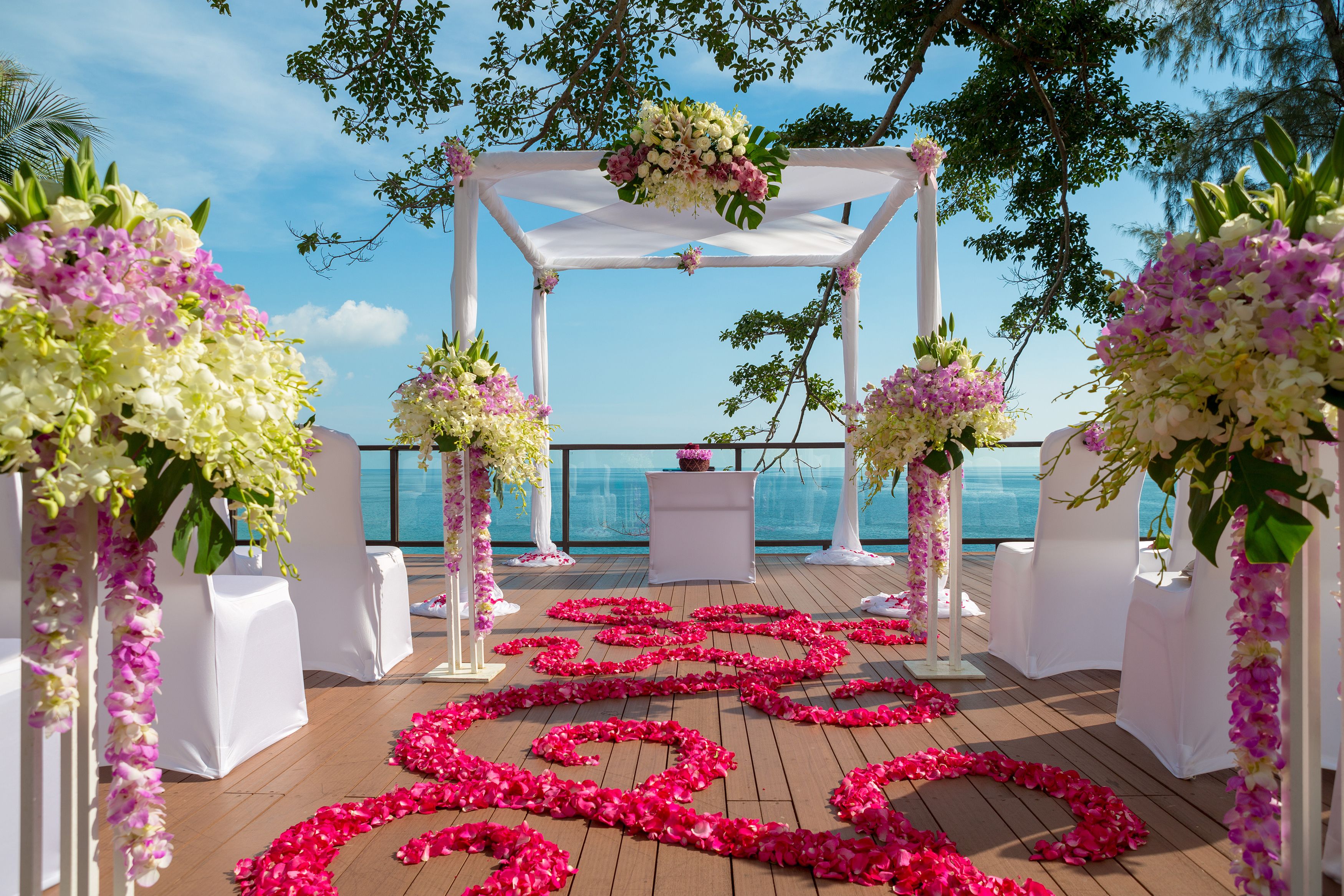 Een buitenbruiloft ceremonie met een houten terras, witte stoelen en bloemendecoraties.